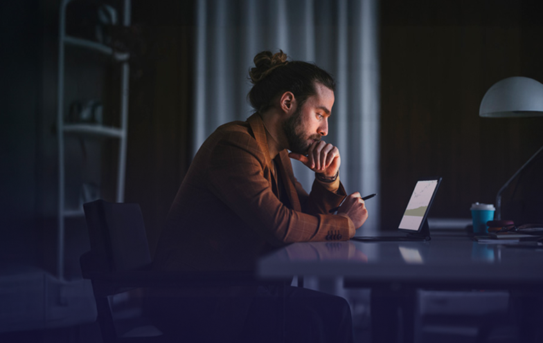 A person with a bun hairstyle works intently on a tablet at a dimly lit desk, with a lamp and a cup nearby.