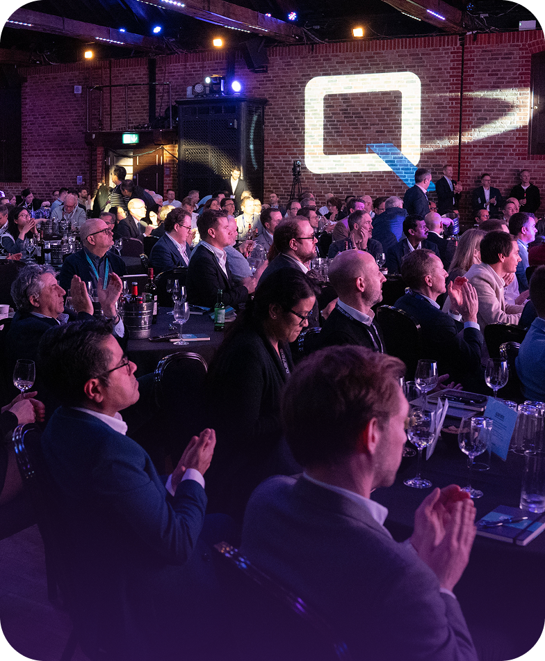 Audience seated at a conference, clapping and attentively watching a presentation in a dimly lit venue with exposed brick walls.