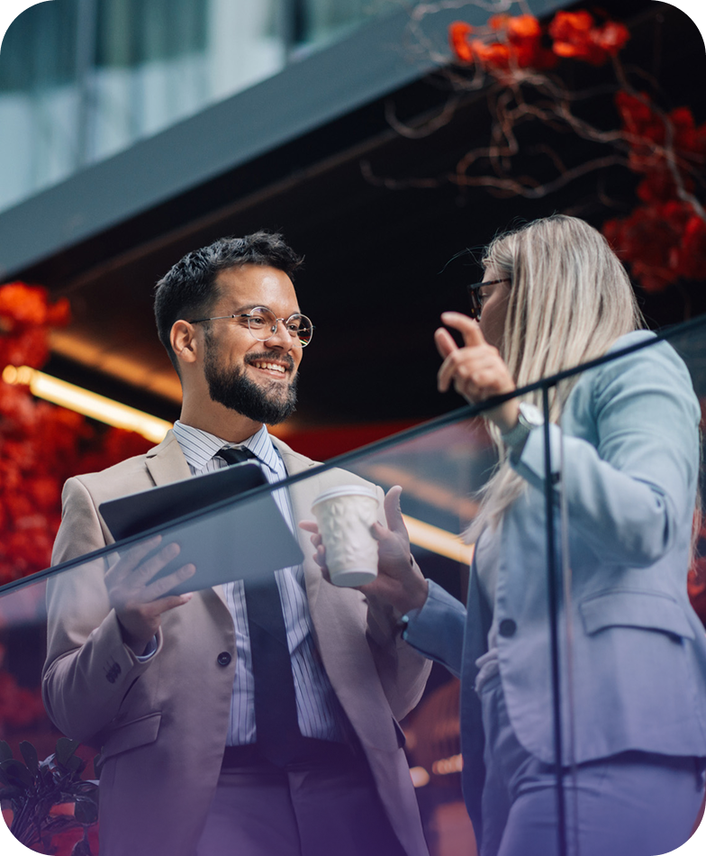 Two professionals in business attire converse cheerfully, one holding a tablet and a coffee cup, in a modern setting with red floral decor.