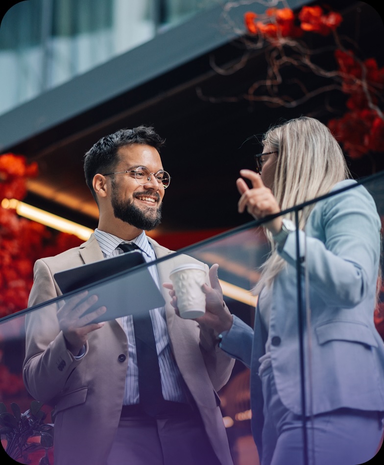 Two professionals in suits converse cheerfully on a balcony, one holding a tablet and coffee cup. Red foliage decorates the background.