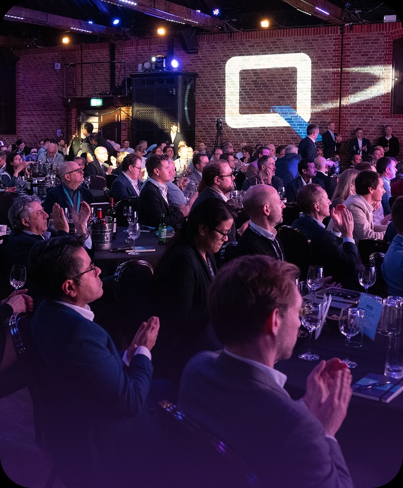 Audience seated at a conference in a dimly lit room with brick walls, clapping and facing a stage with a projected logo.