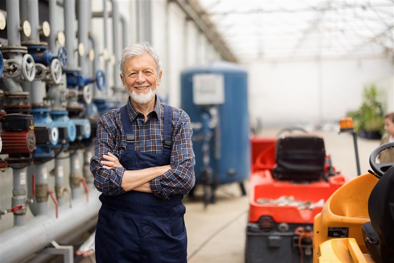 Elderly man with crossed arms, wearing overalls, stands in an industrial workshop with machinery and pipes in the background.