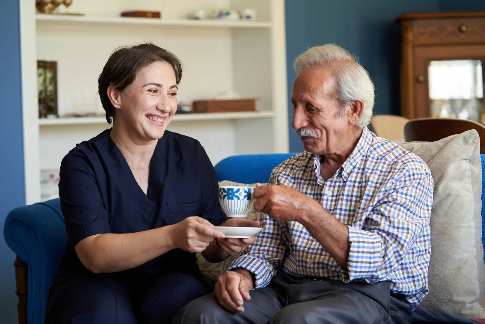 A woman with short hair hands a teacup to a smiling elderly man in a cozy living room setting.