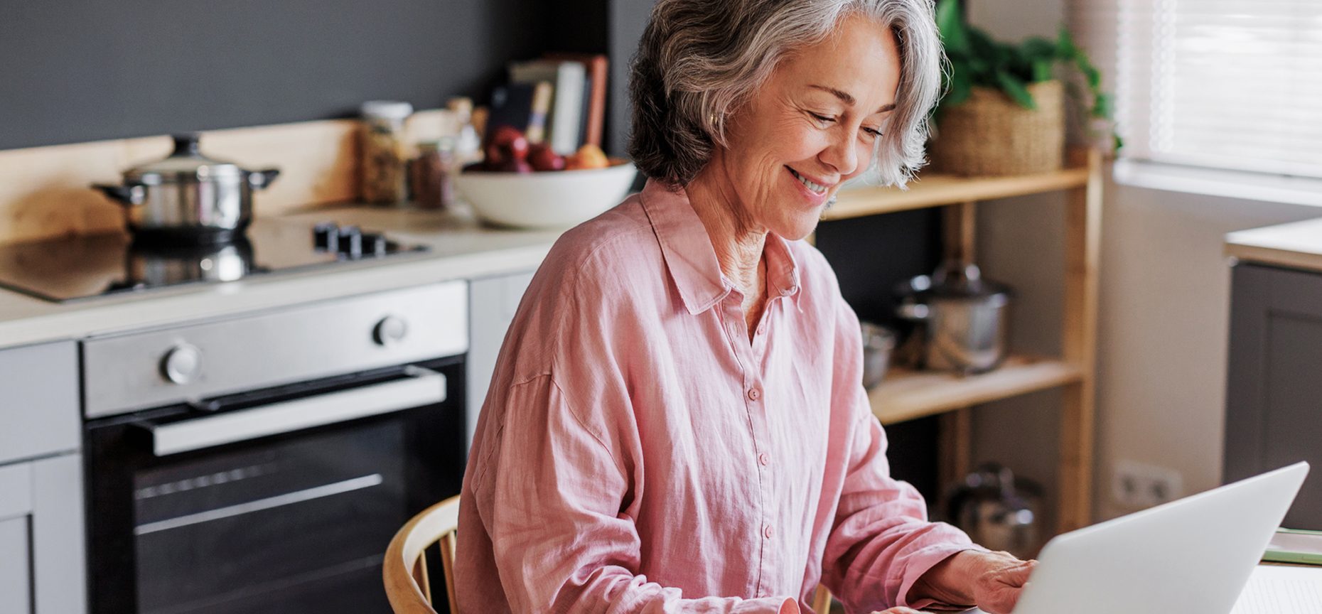 Smiling woman with gray hair using a laptop in a cozy kitchen, wearing a pink shirt. Stove, cookware, and shelves visible in the background.
