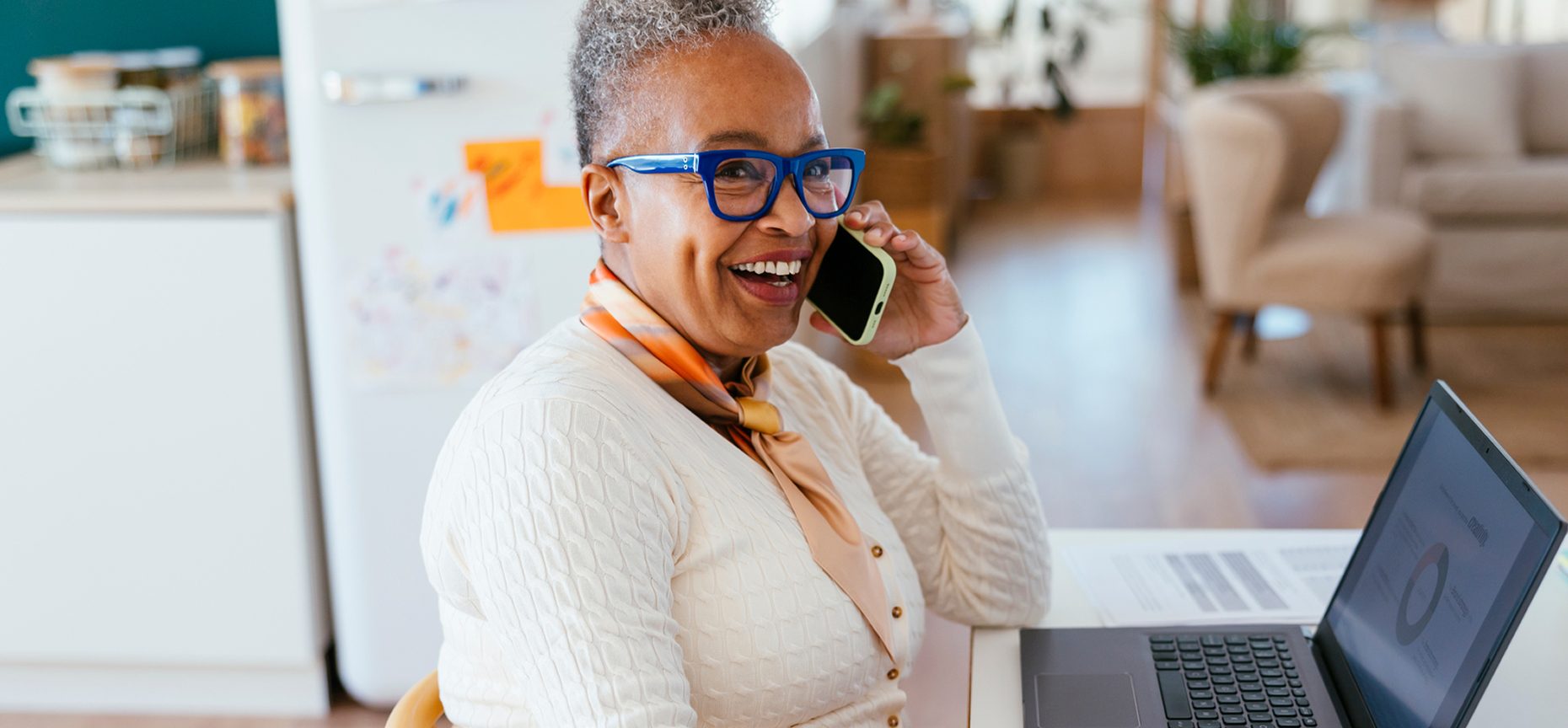 Elderly woman with blue glasses, smiling and talking on the phone, sits at a table with a laptop displaying a pie chart.