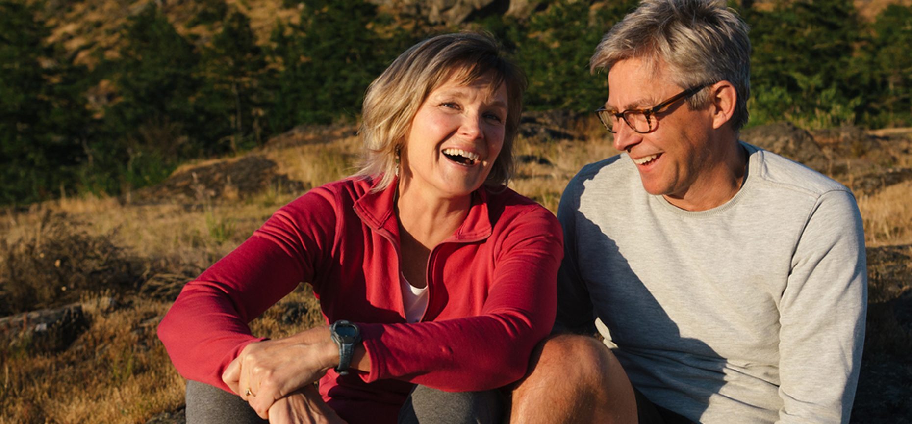 A smiling couple sits together outdoors in casual clothes, enjoying a sunny day with trees and hills in the background.