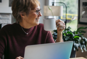 A woman with short hair, wearing glasses, smiles while holding a mug and sitting at a table with a laptop. A plant and window are in the background.