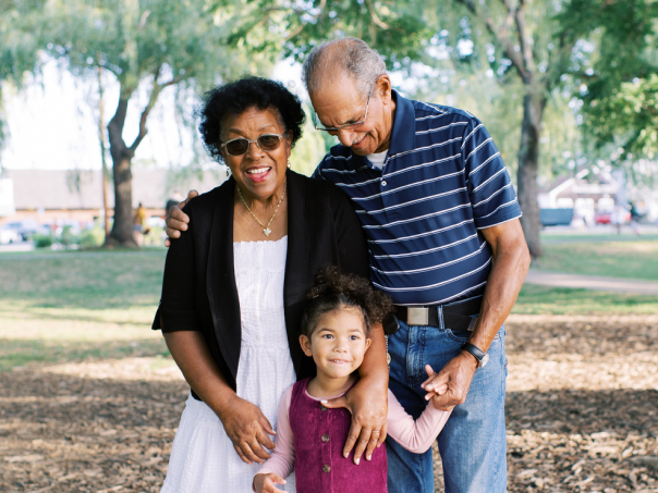 A smiling elderly couple with a young girl standing between them, outdoors in a park on a sunny day.