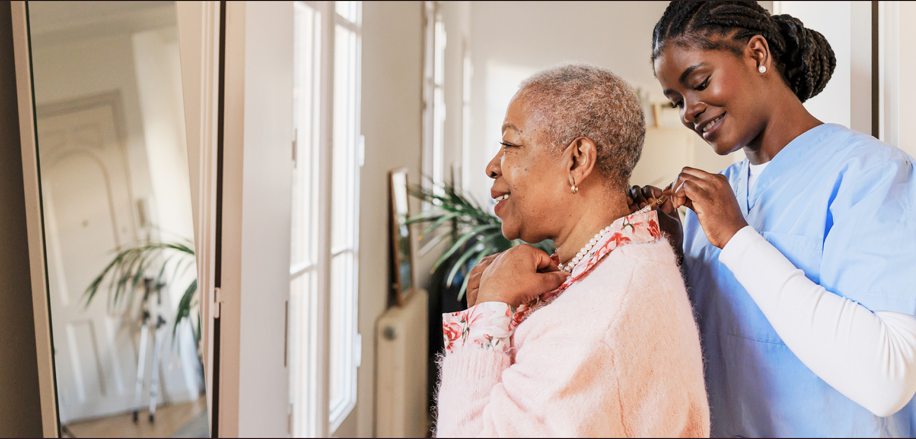 Caregiver in blue scrubs helps elderly woman fix her necklace, both smiling warmly in a bright, cozy room.