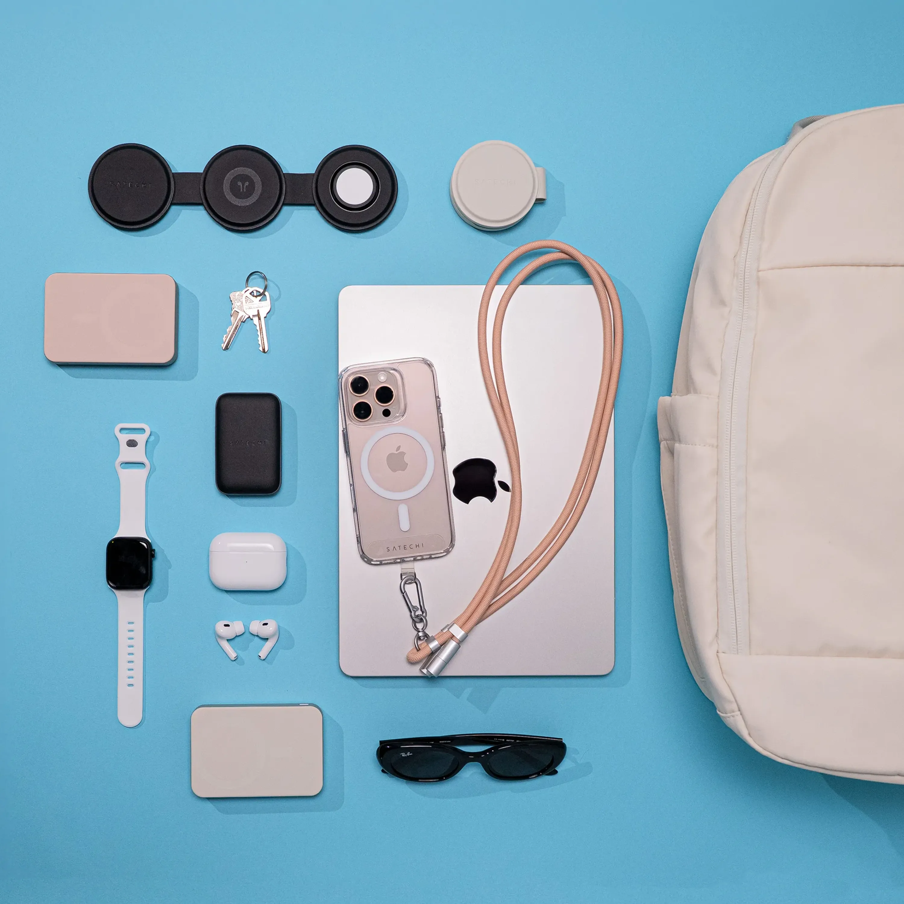 Flat lay of Satechi products on a blue background, featuring OntheGo™ USB-C Lanyard Cable, OntheGo™ 67W Slim Wall Charger, OntheGo™ 3-in-1 Charger, a Apple Watch, AirPods, iPhone with clear case, MacBook, charging cables, keys, sunglasses, and a beige backpack.