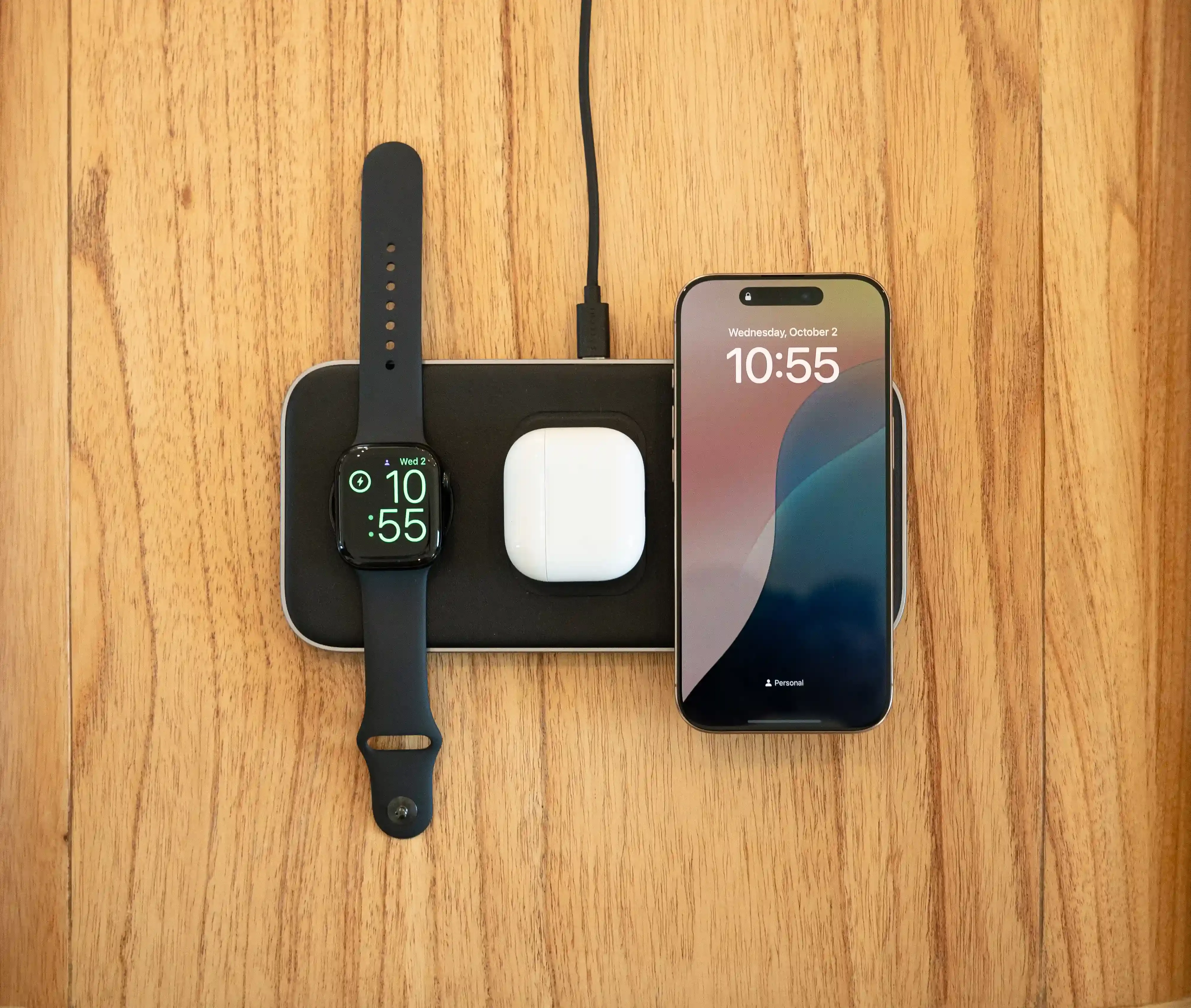 A Qi2 Trio Wireless Charging Pad charging an iPhone, AirPods, and Apple Watch on a wooden side table.