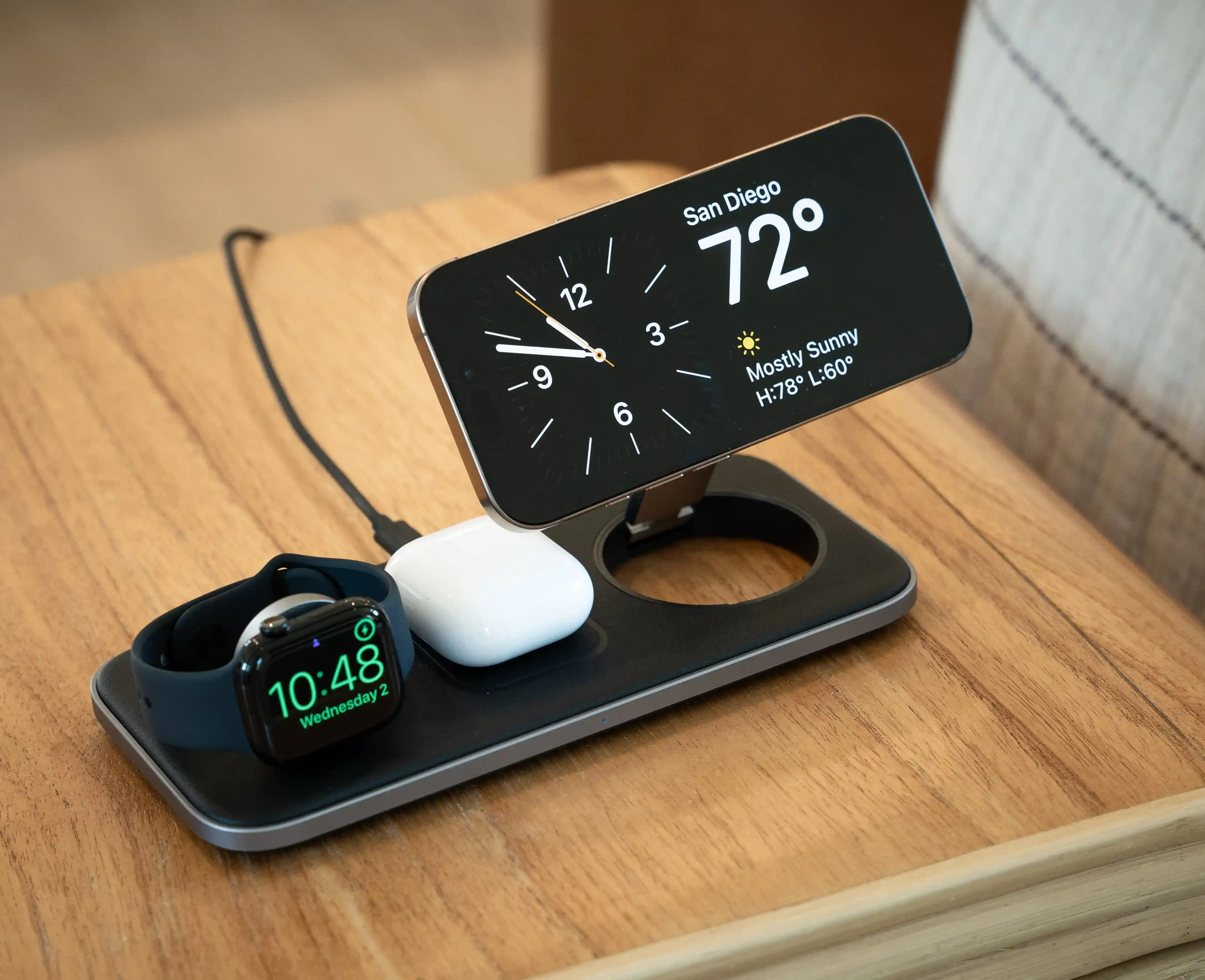 A Qi2 Trio Wireless Charging Pad charging an iPhone, AirPods, and Apple Watch on a wooden side table.