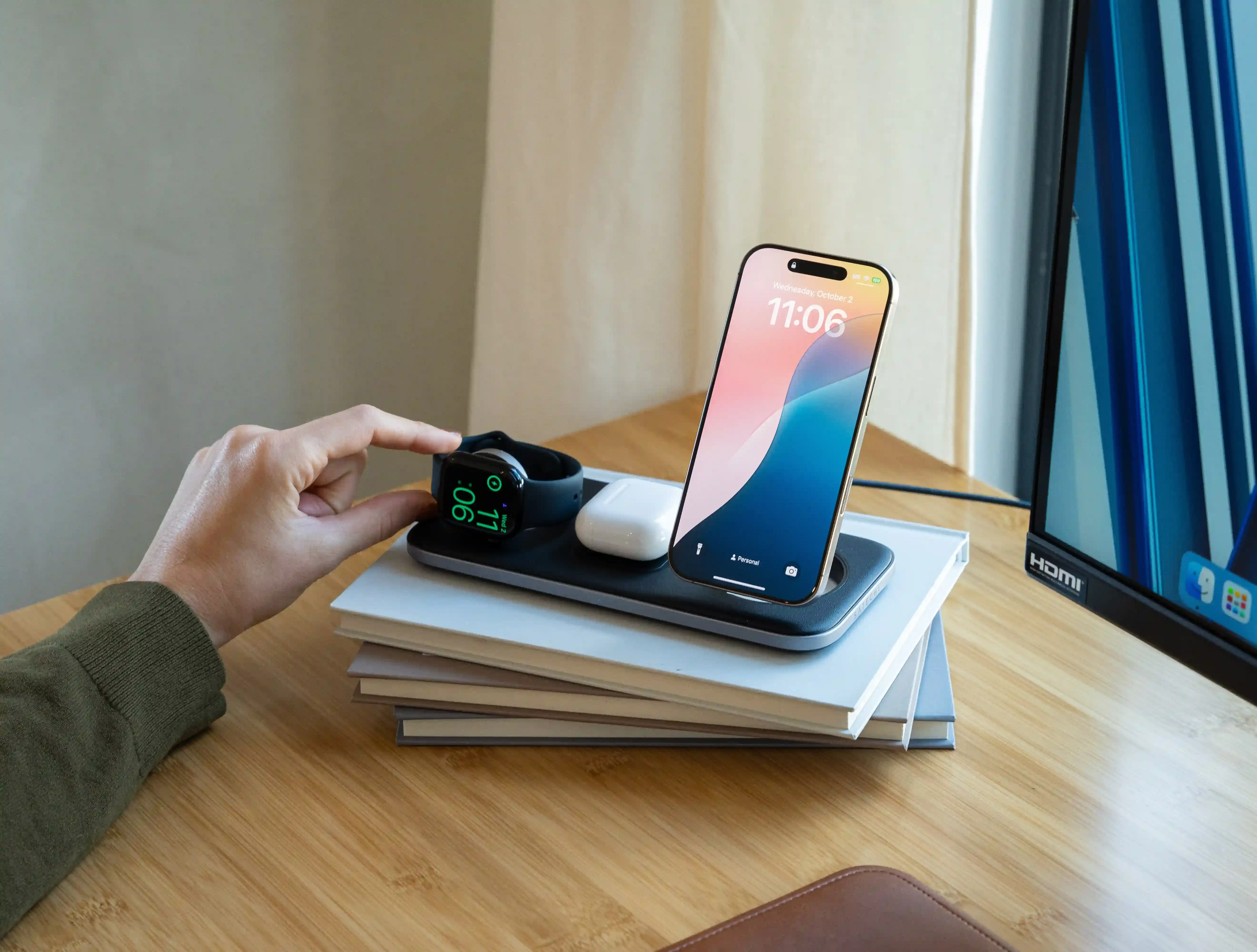 A person’s hand adjusting a sleek Qi2 Trio Wireless Charging Pad, simultaneously charging an iPhone, AirPods, and an Apple Watch, all resting on a neatly stacked pile of hardcover books.