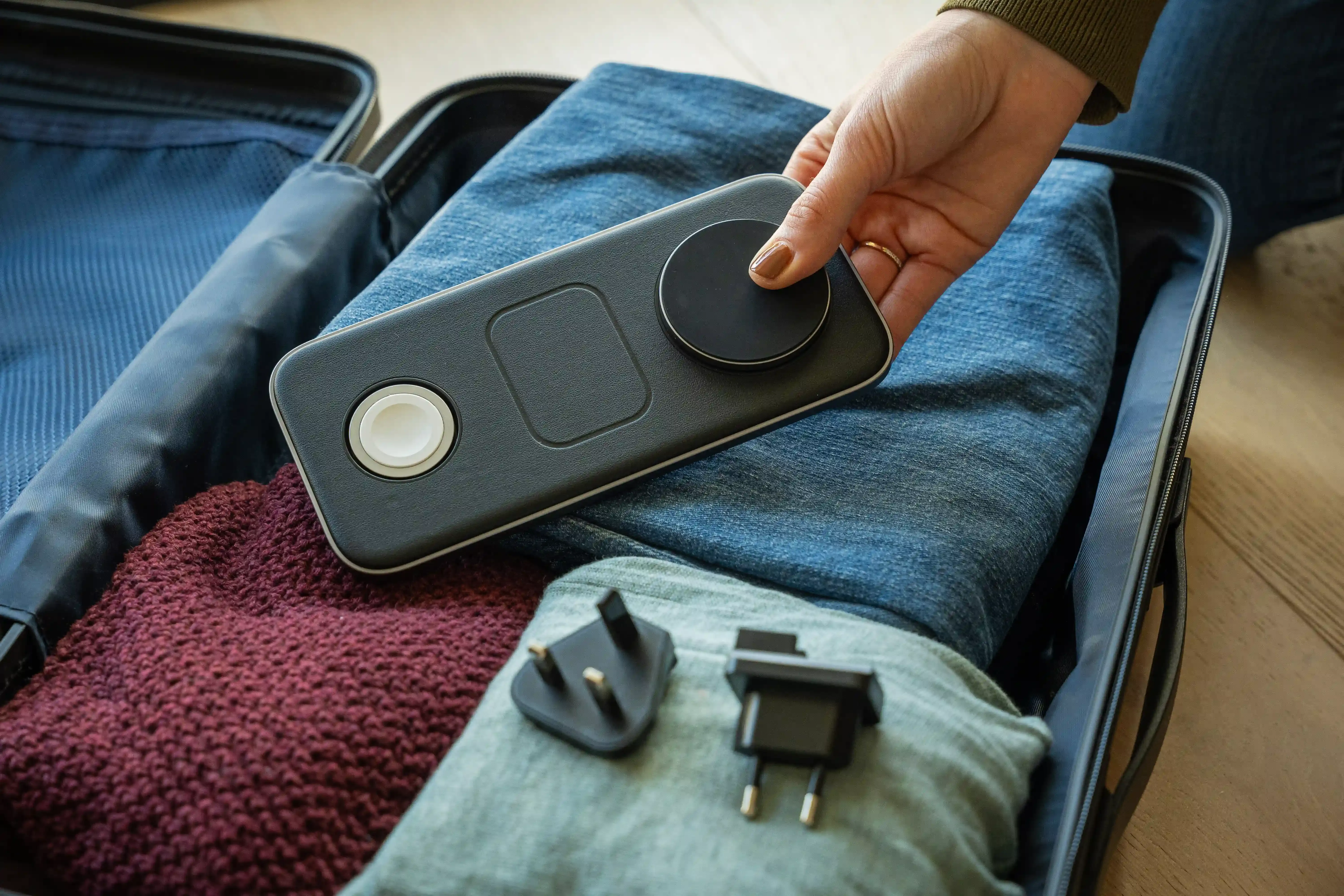A hand placing a Qi2 Trio Wireless Charging Pad into a suitcase filled with folded clothes.