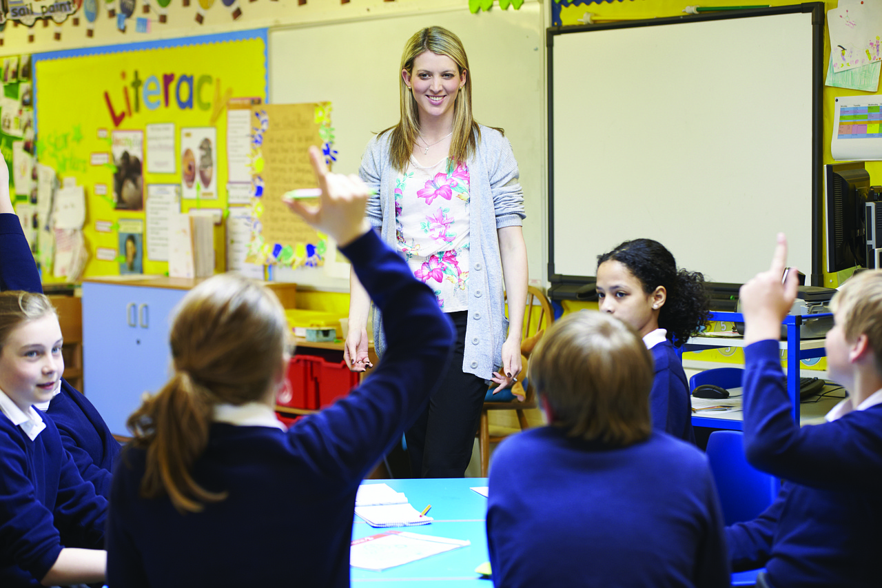 A teacher in a classroom, in front of her young students. One of the students has her hand raised.