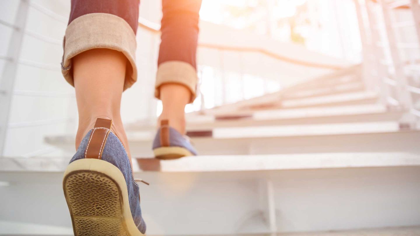 The feet of a woman wearing blue cloth sneakers walking up a flight of stairs into the sunlight.