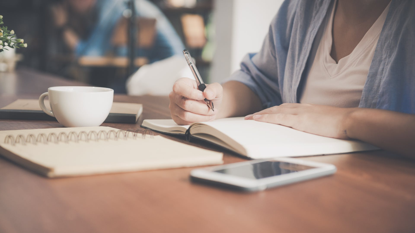 Someone's hands as they write with a pen in a notebook on a desk with other study materials