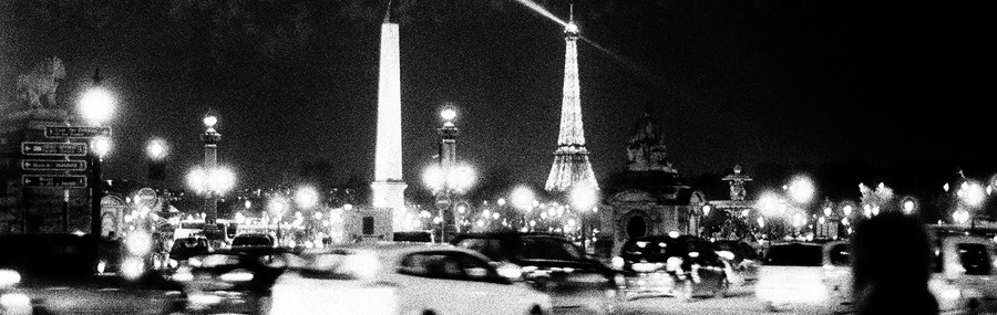 A night shot of the Eiffel Tower in the background with cars and people on the street