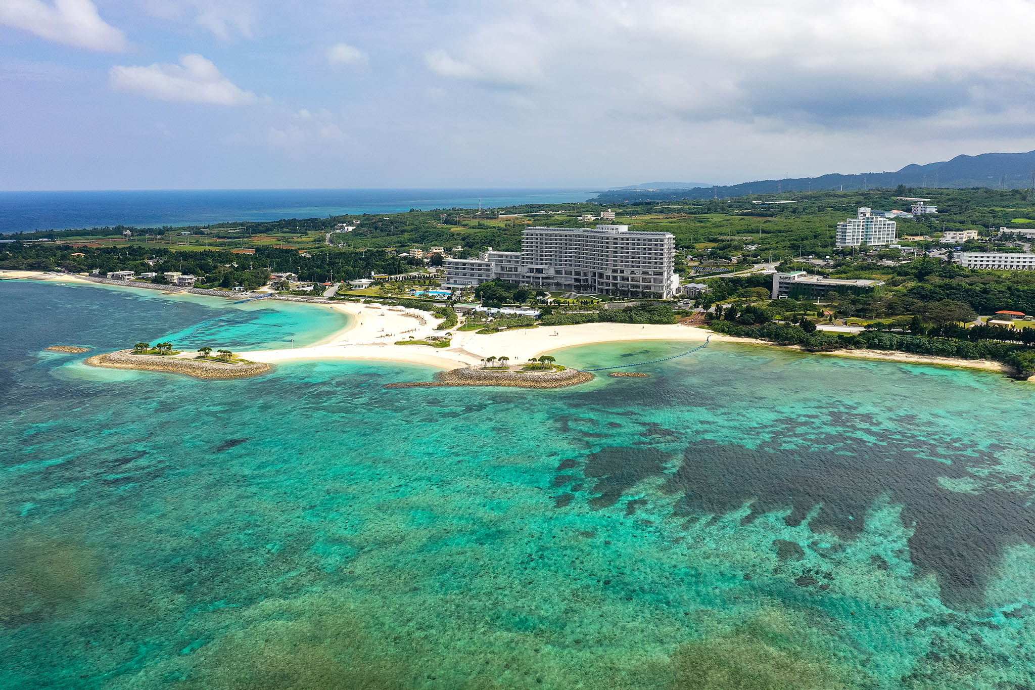 An aerial shot of the clear turquoise waters off the coast of Okinawa. There are some large buildings, but most of the landscape is green and healthy.