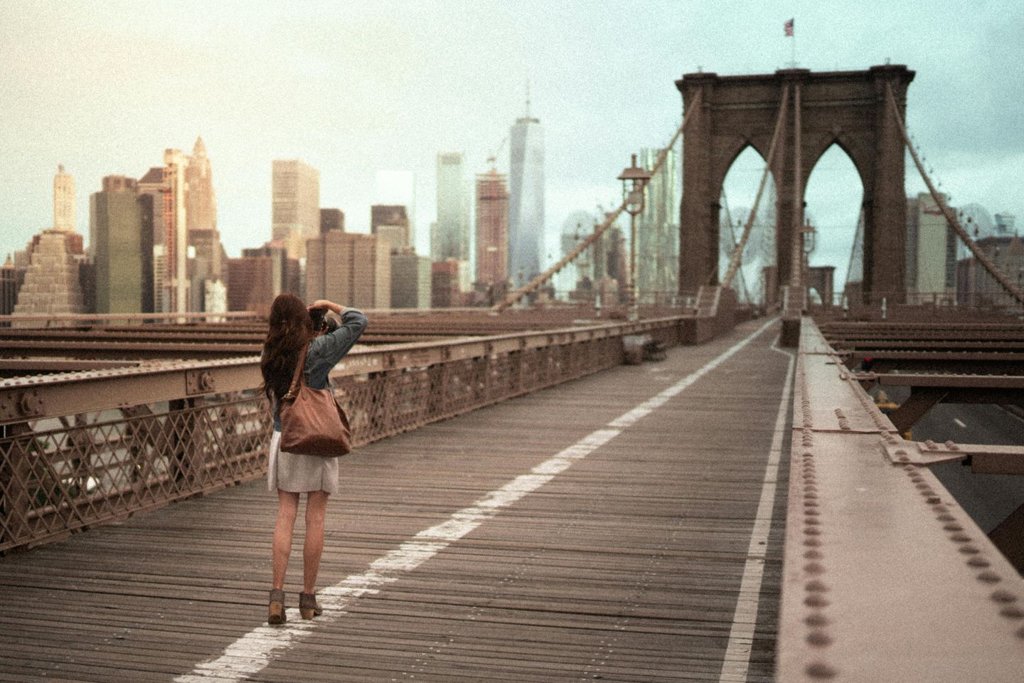 A tourist walking the Brooklyn bridge taking a picture of the New York City skyline