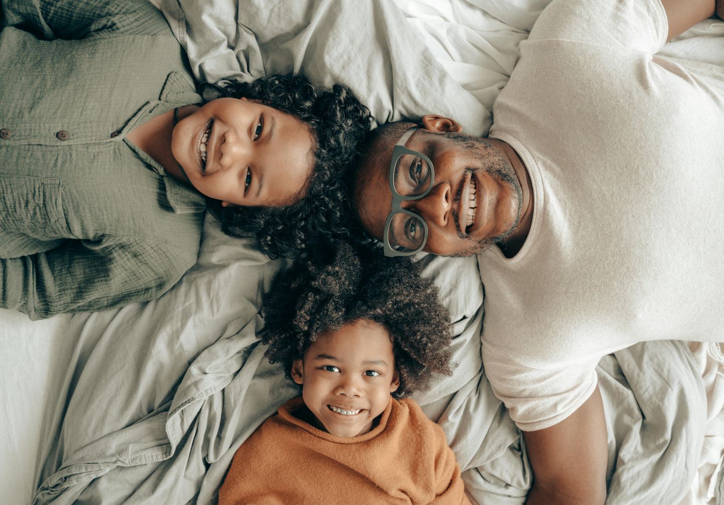 A photo taken from above of a father and two children lying on a bed with their heads together in a triangle. The father is a black man in glasses and the children both look younger than 10 and have curly hair.