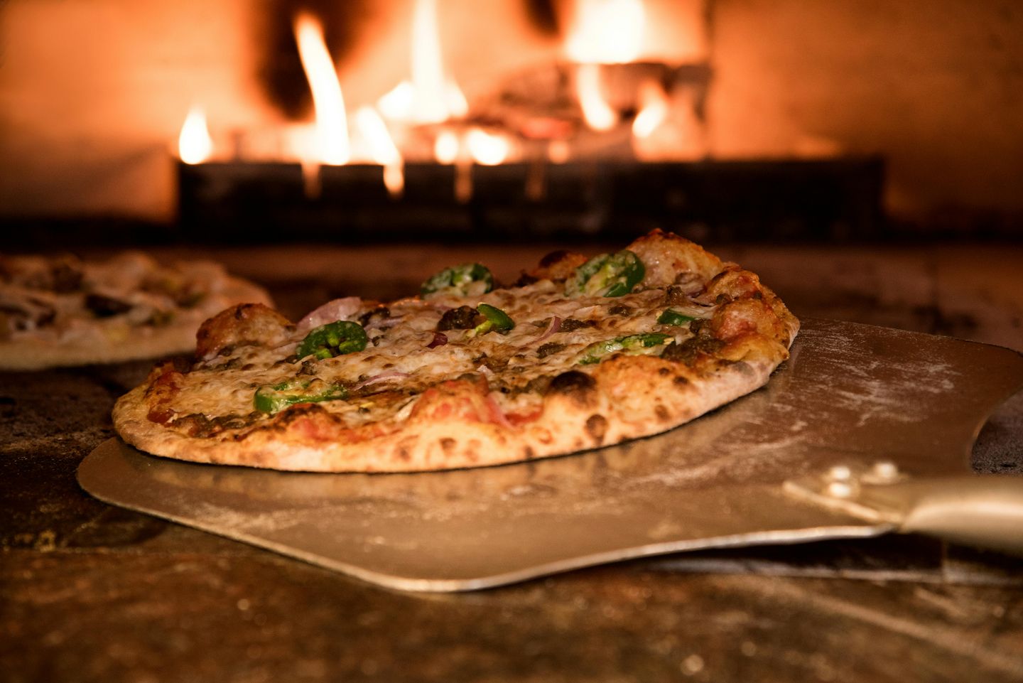 A pizza being put inside a wood oven with a lit fire in the background