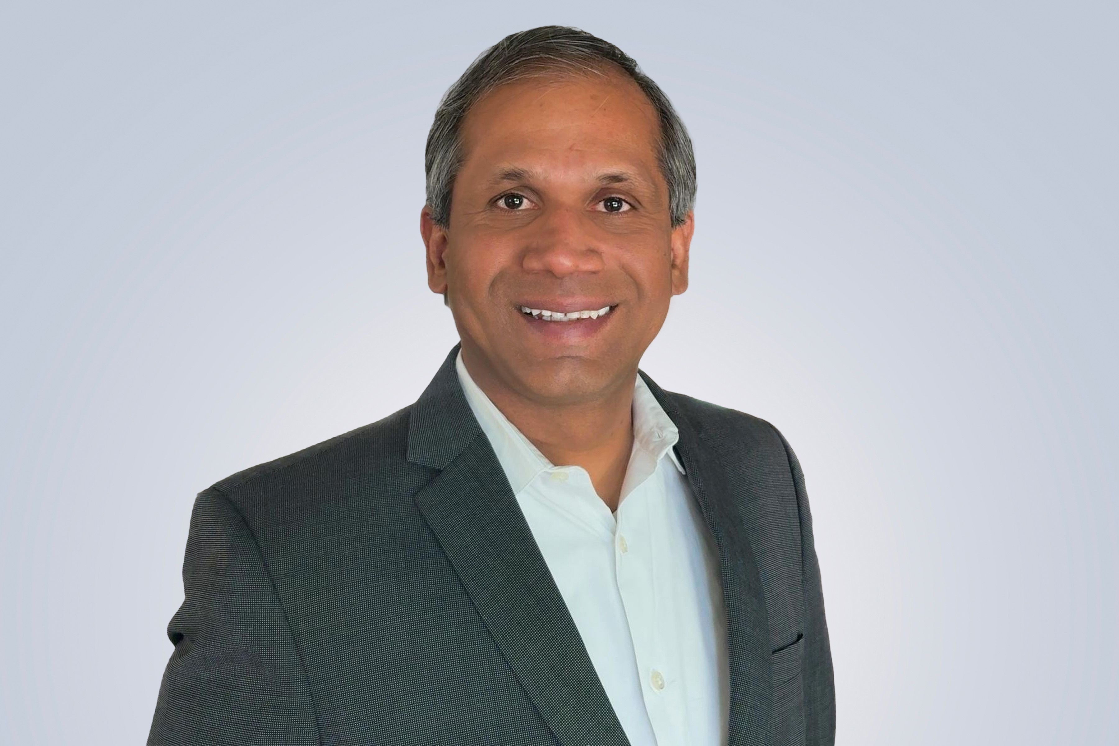 Smiling man in a dark suit and white shirt against a light gray background.
