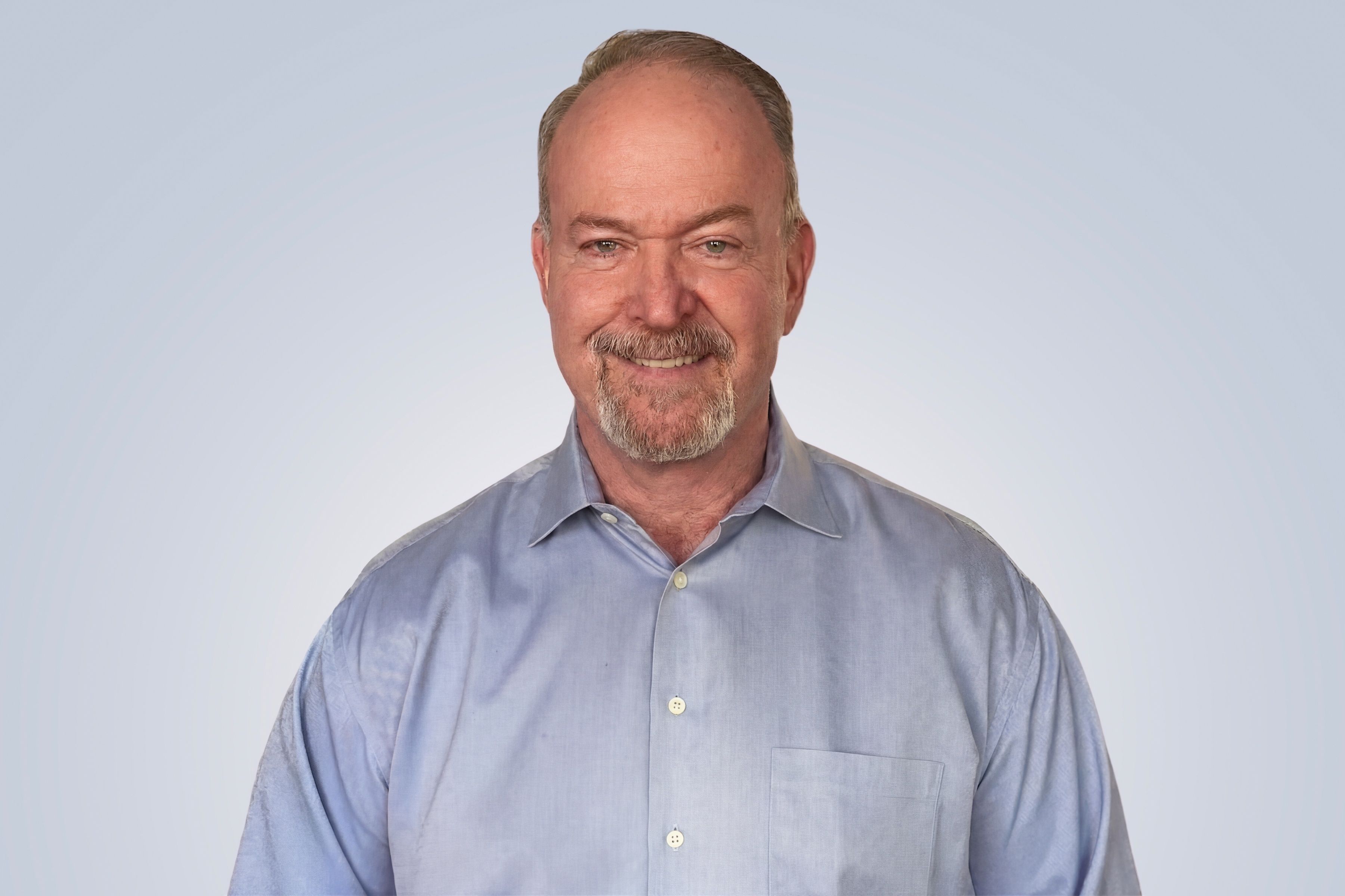 Smiling man with a beard wearing a light blue button-up shirt against a light gray background.