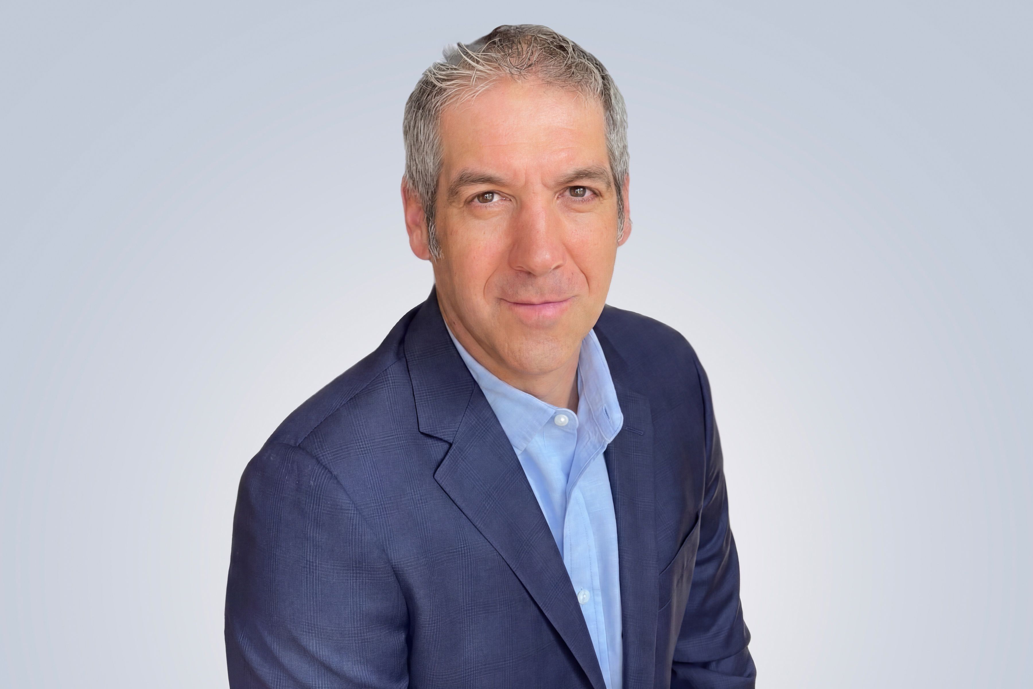 A man with short gray hair wearing a blue blazer and light blue shirt, posing against a light gray background.