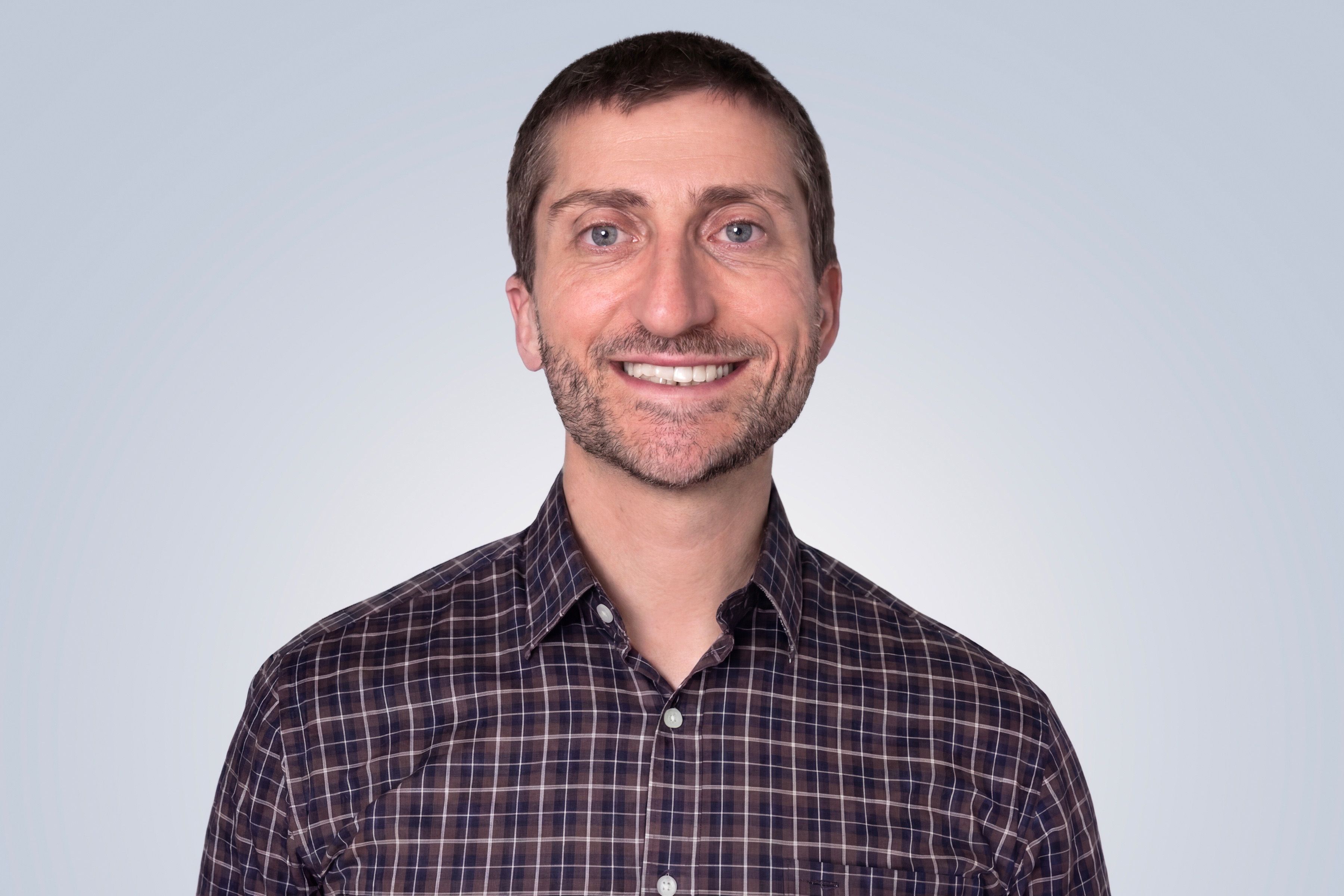 Smiling man with short hair in a plaid shirt against a light gray background.