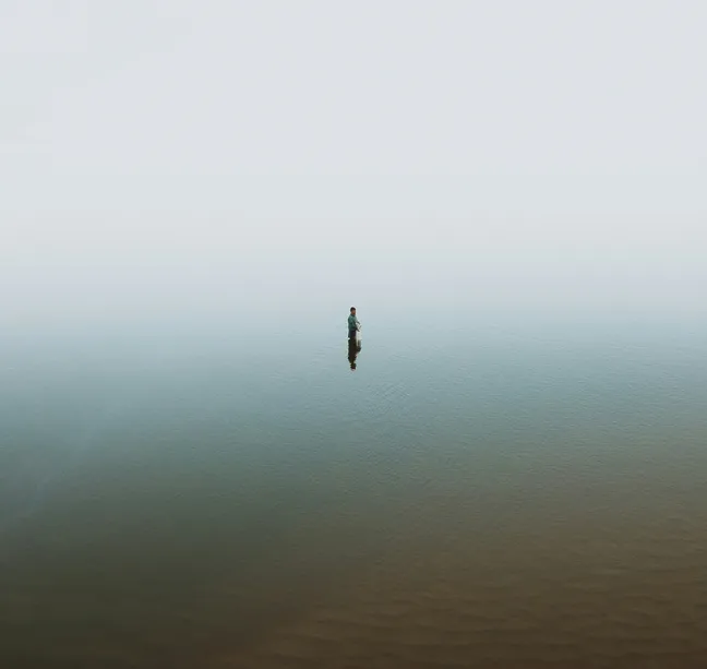 A lone fisherman stands waist-deep in a misty body of water during daytime, surrounded by fog, creating a serene and isolated atmosphere