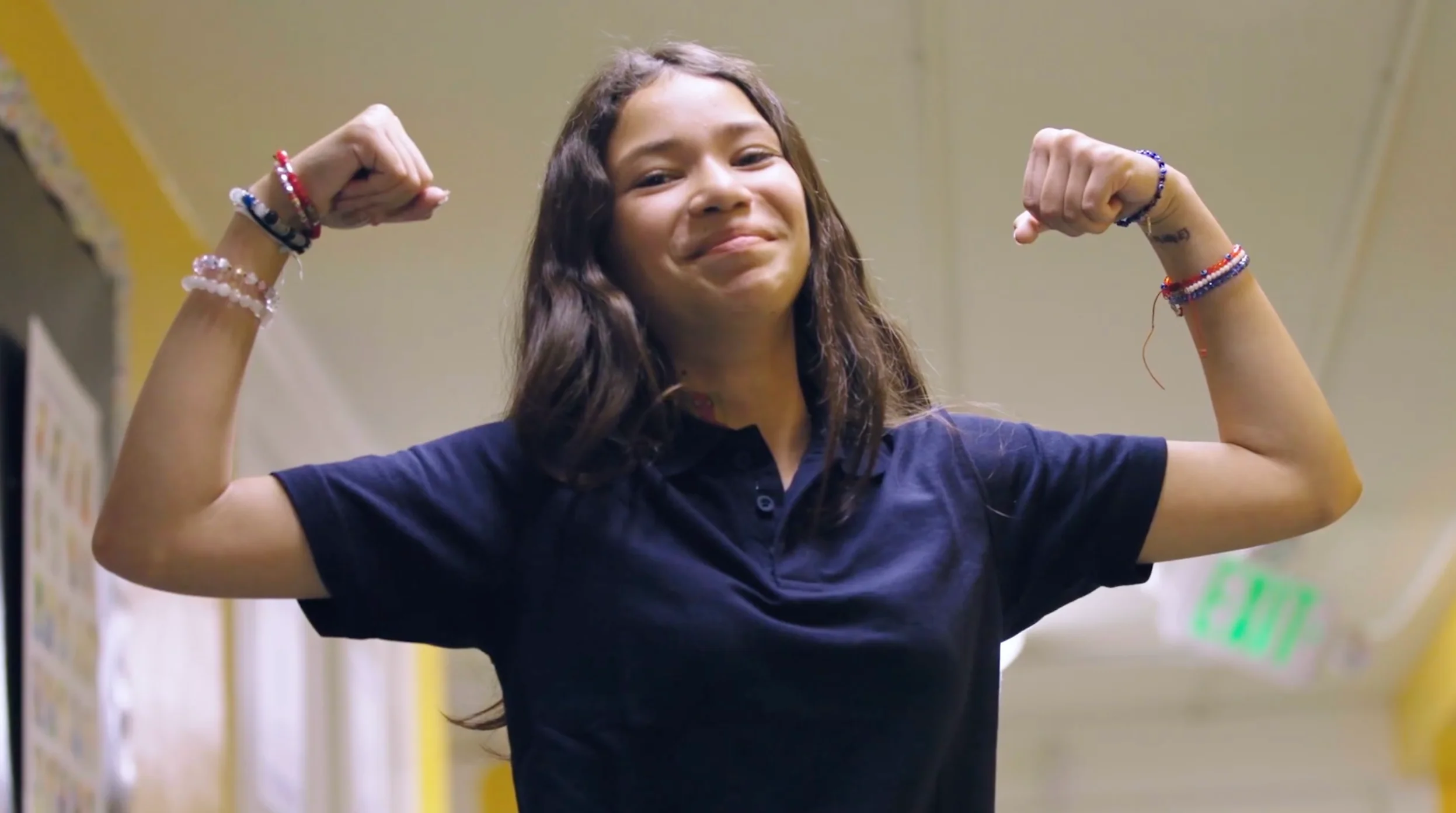 Girl in a navy shirt smiling and flexing her arms, wearing colorful bracelets, stands in a hallway.