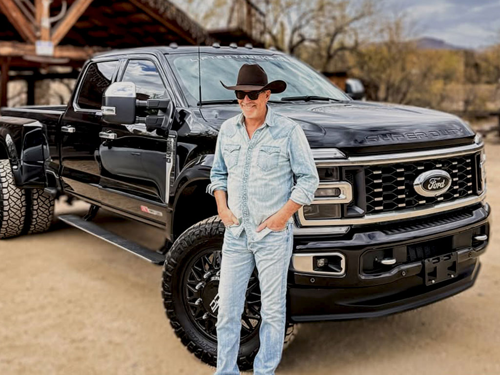 Kevin Costner leaning against a big lifted Ford F-450 Dually smiling