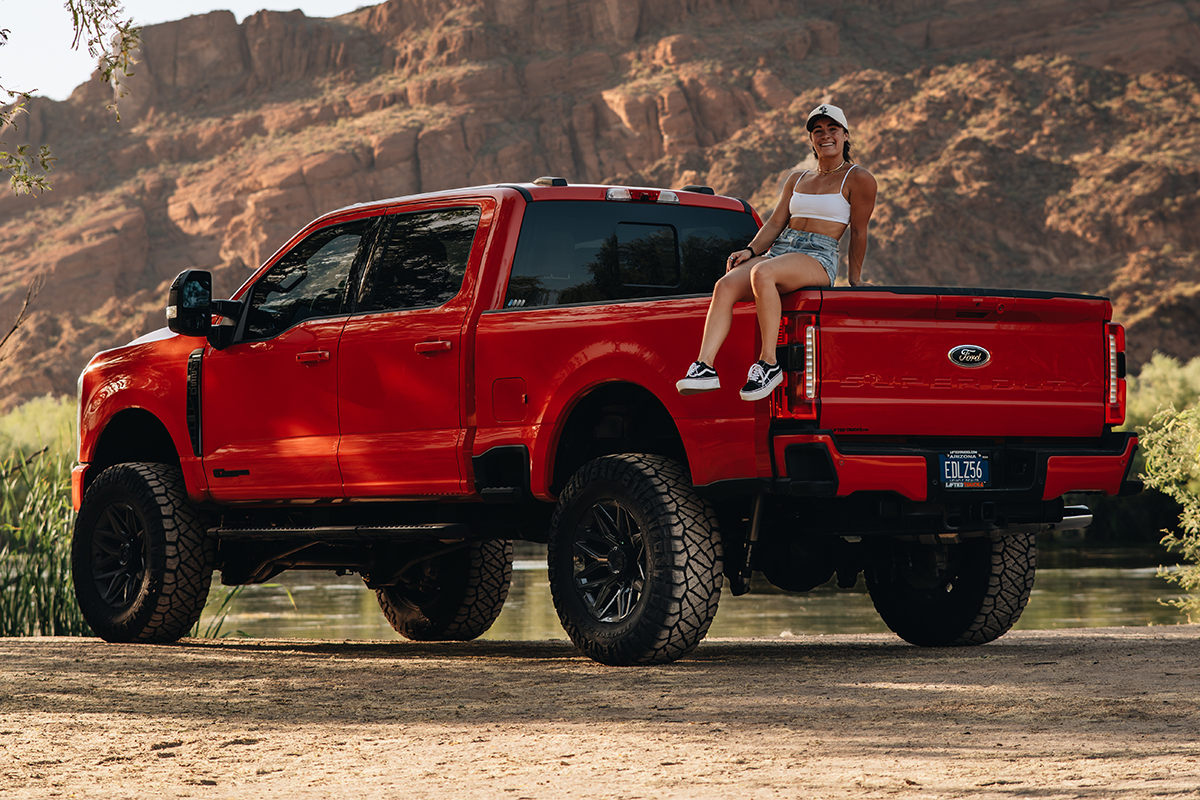 young lady in a white tank top and shorts sits on the bed of a red Ford super duty that has been lifted and has black rims.