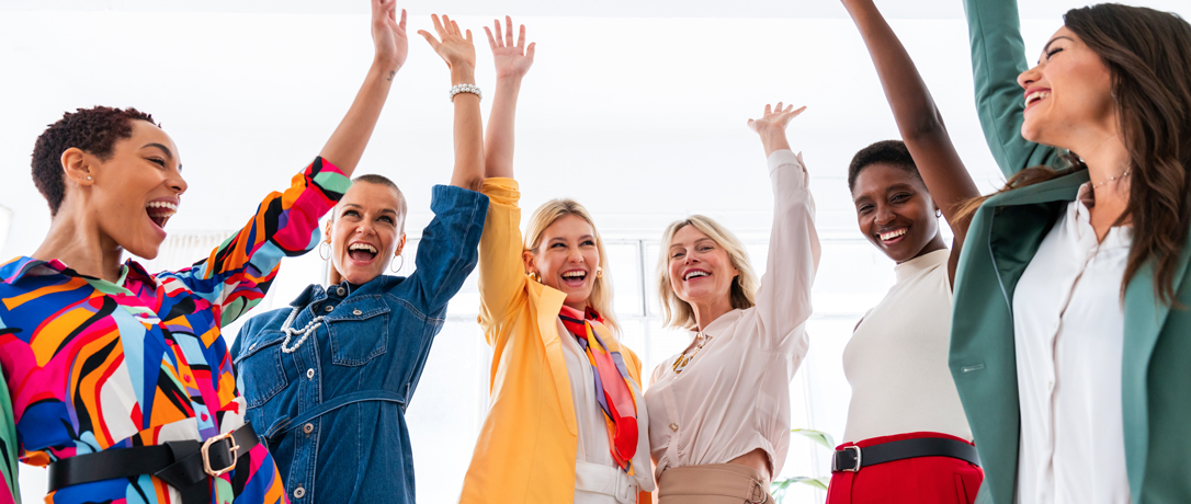 A group of six diverse women joyfully raising their arms in celebration while smiling, dressed in colorful, stylish outfits in a bright room.