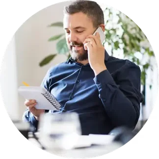 Man in a blue shirt talking on the phone, holding a notebook with a pencil, sitting at a desk with blurred greenery in the background.