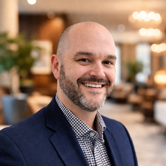 Smiling man in a blue blazer and checkered shirt, standing in a warmly lit modern interior with soft-focus lights and plants in the background.