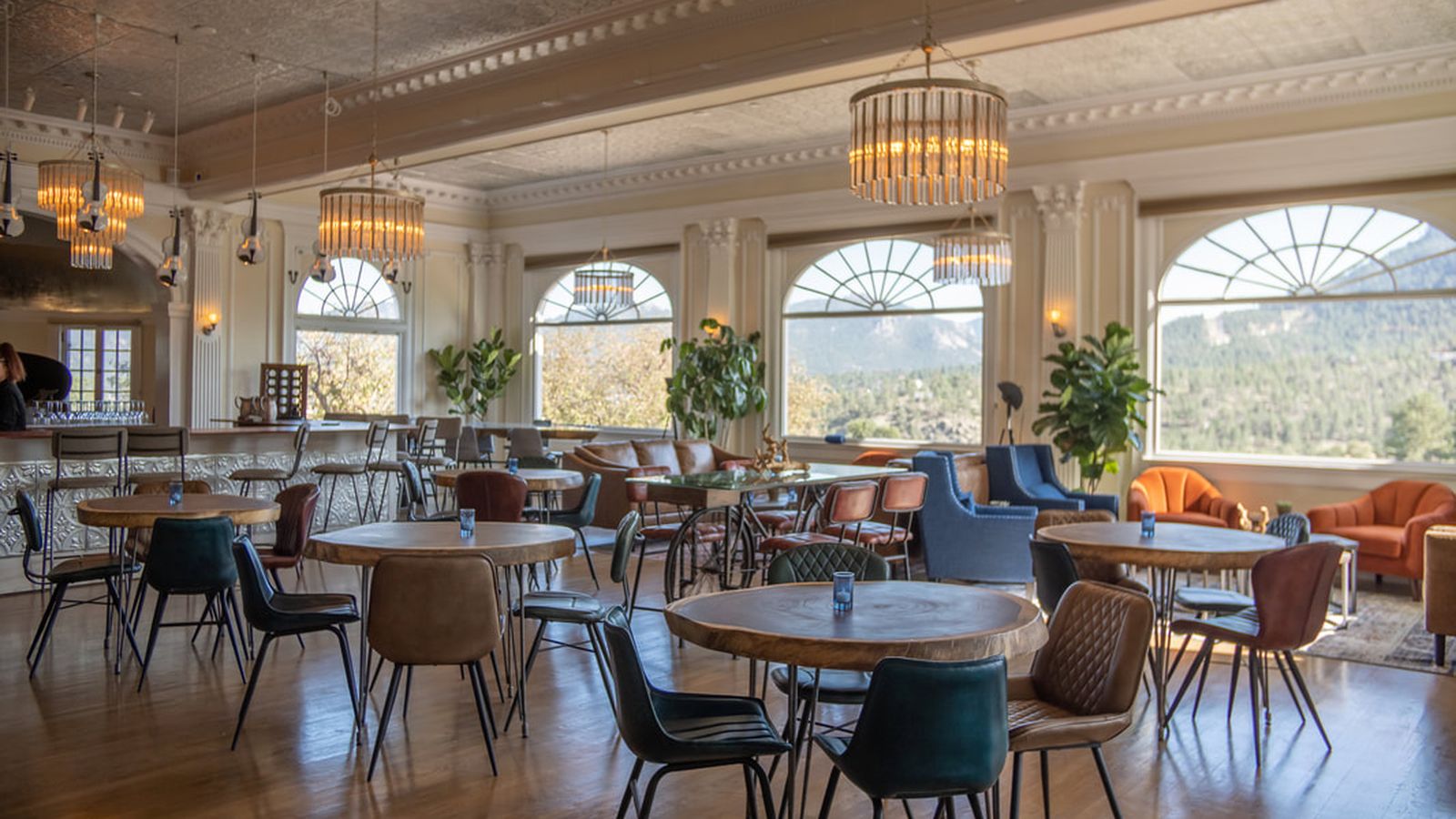 Spacious, elegant café with round wooden tables, diverse chairs, large windows, chandeliers, and mountain views.