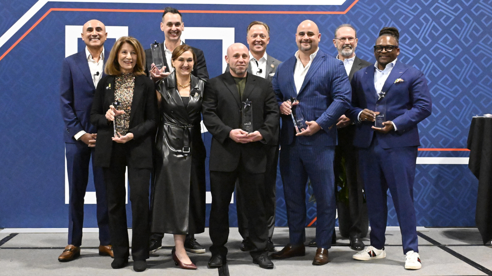 Nine people in formal attire stand in a row, holding awards, against a blue patterned backdrop.