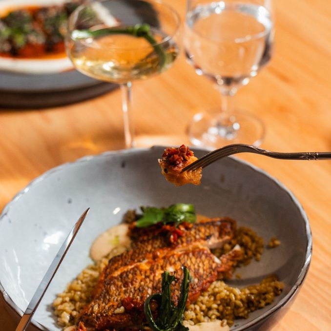 A person holds a fork with food over a dish of seasoned fish and grains. Glasses of drinks and a pizza are on the wooden table.