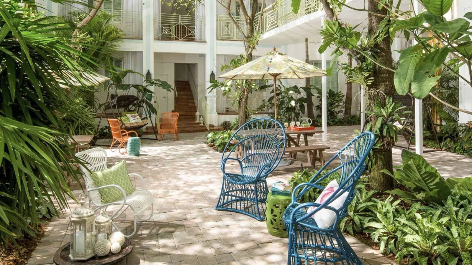 Lush courtyard with blue wicker chairs, a picnic table, and tropical plants. White building with balconies in the background.