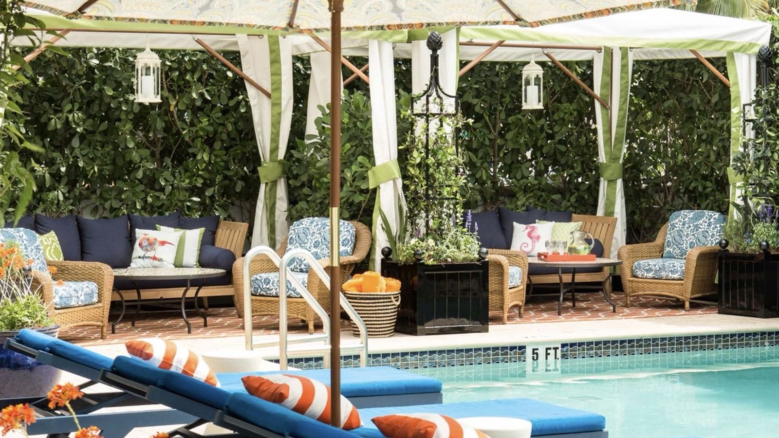 Poolside scene with wicker chairs, colorful cushions, and striped loungers under umbrellas. Lush greenery and lanterns enhance the cozy atmosphere.