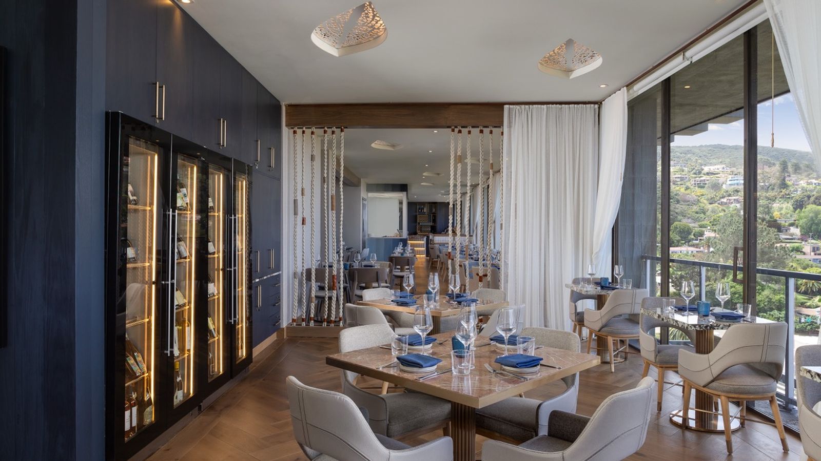 Elegant restaurant interior with modern decor, featuring wooden tables, gray chairs, large windows, and a scenic view outside.