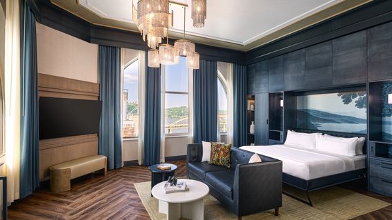 Luxurious hotel room with a chandelier, king bed, leather sofa, wooden floor, and large windows with blue curtains offering a scenic view.