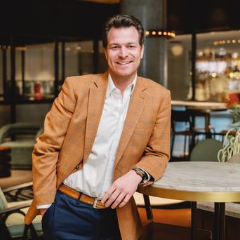 Man in an orange plaid blazer leans on a marble table, smiling in a stylish, modern interior with soft lighting.