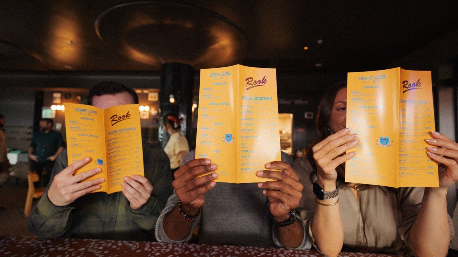 Three people sitting at a table in a dimly lit restaurant, each holding a yellow menu that covers their faces.