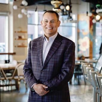 A person in a plaid suit smiles in a modern cafe with colorful decor and empty seating.