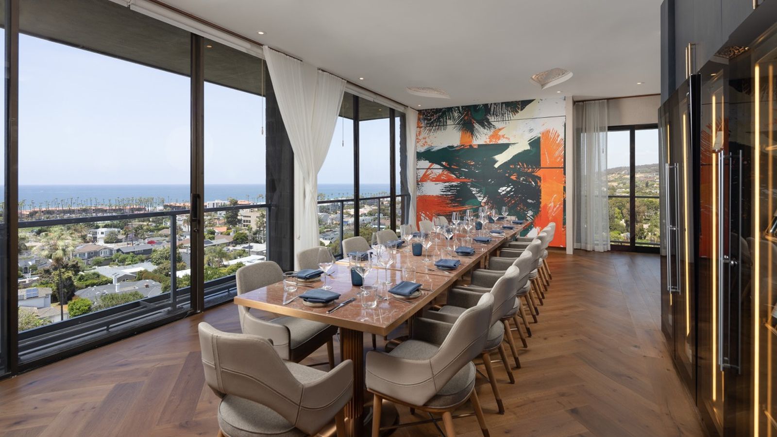 Modern dining room with a long table set for a meal, surrounded by chairs. Large windows offer a scenic view of the ocean and cityscape.