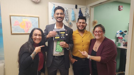 Four people smiling and pointing at a man holding a book and socks in an office decorated with a fish painting and wall hangings.