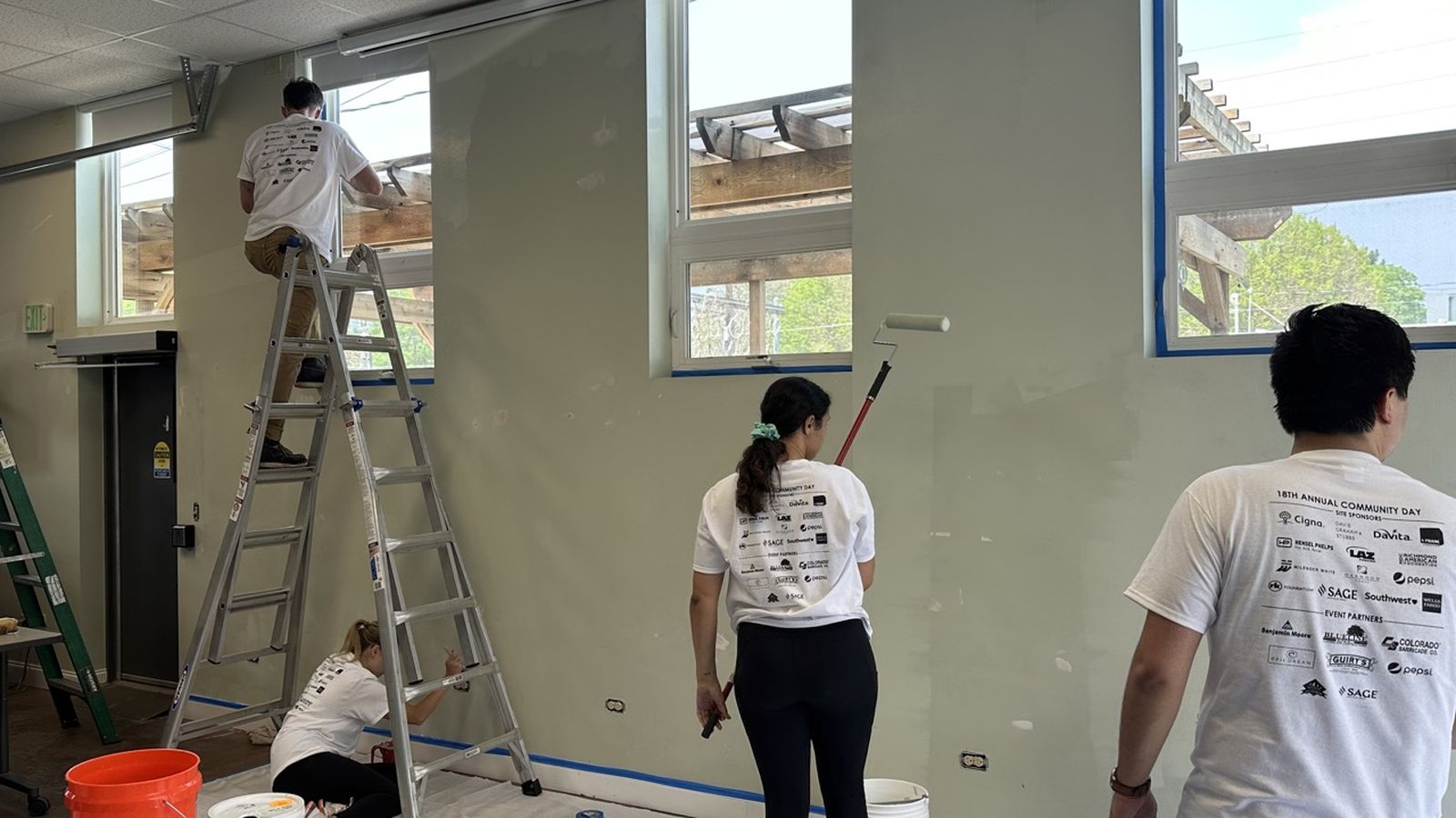 Volunteers painting a room with light green walls; one on a ladder, others using rollers and brushes, wearing matching white shirts.